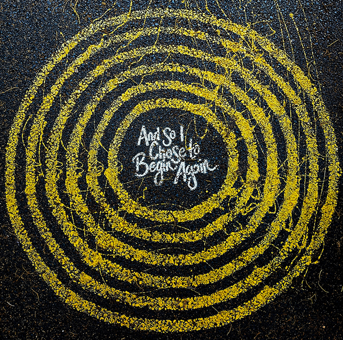 Painted concentric yellow circles on black pavement. In the center circle, white letters state: And So I Chose to Begin Again